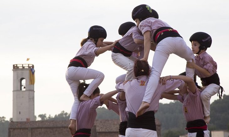 Humans in white clothing with black helmets climbing on top of each other to form a human structure with a church tower in the background.